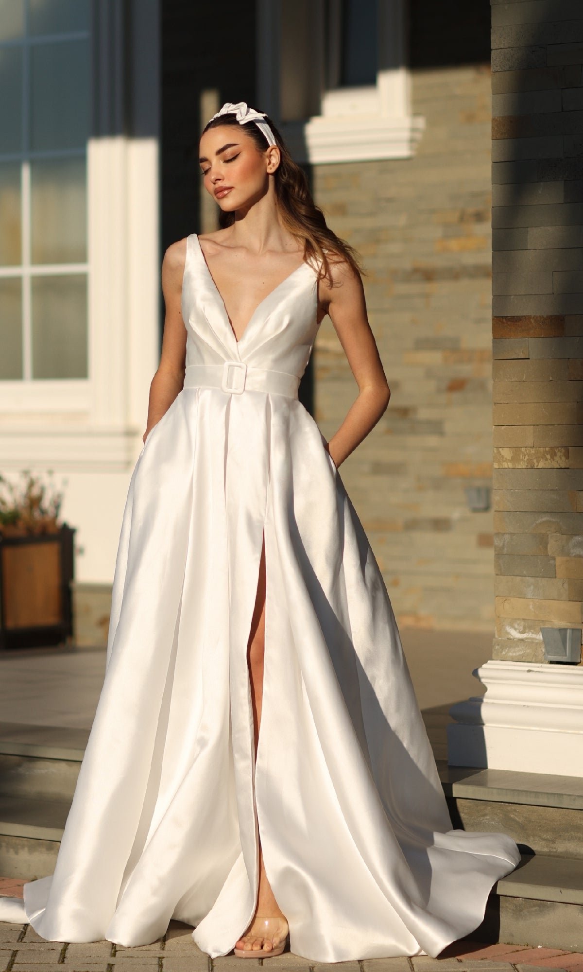 A woman stands outdoors in the Tina Holly BB209W Belted Long White Prom Dress, featuring a deep V-neck, belted waist, and high front slit. She pairs it with a headband near a stone building, radiating elegance in this formal gown.
